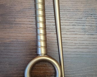 May include: A gold-colored metal tool with a ring and a long, thin rod. The handle of the ringed part has a spiral design. The rod has a black tip. The items are on a dark wood surface.