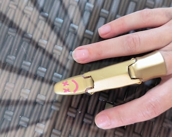 Puede incluir: Una armadura de dedo dorada con un diseño articulado. La punta de la armadura presenta una superficie amarilla con una carita sonriente rosa. La armadura está diseñada para cubrir el dedo y el nudillo.