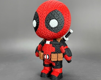 Puede incluir: Figura de Deadpool roja y negra con apariencia de punto texturizado. La figura tiene grandes ojos negros con detalles blancos, un cinturón de herramientas y una espada en la espalda. La figura está sobre una superficie gris.