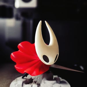 Może przedstawiać: Wydrukowana w 3D figurka postaci z białą głową i czerwonym płaszczem. Figurka jest zamontowana na białej, teksturowanej podstawie. Tło jest rozmyte, ze skupieniem na figurce.