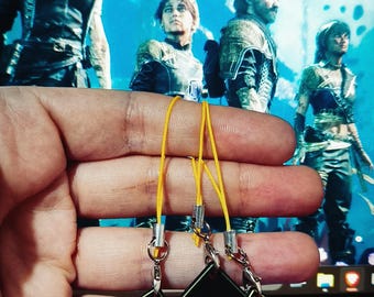 Puede incluir: Tres llaveros negros en forma de diamante con el número "33" en dorado. Cada llavero tiene un cordón amarillo. El fondo presenta una escena con varias personas vestidas con ropa futurista.
