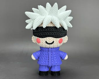 May include: A handmade figurine of a character with spiky white hair, a black blindfold, and a blue outfit. The figure has rosy cheeks and a small smile. The doll is made of a textured material, possibly yarn or fabric.