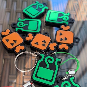 Peut inclure: Une collection de porte-clés et de breloques vertes et oranges. Les breloques ont un contour noir et présentent une forme de bouteille ou de bonbon. Certains ont des porte-clés et des cordons colorés attachés.