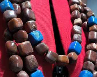 May include: A layered necklace featuring dark brown wooden beads interspersed with vibrant blue square beads. The necklace is displayed on a red dress form, showcasing its design and color contrast.