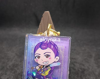May include: A rectangular keychain featuring a cartoon character with purple hair and a yellow and purple outfit. The keychain has a clear border with the text "aloynika" at the top. The background is a dark surface.