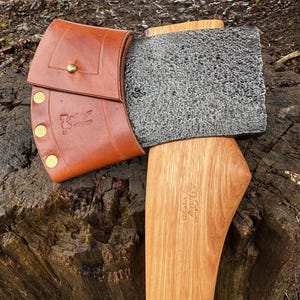 Könnte beinhalten: Eine handgefertigte Axt mit Holzgriff und strukturiertem Stahlkopf. Die Axt hat eine braune Lederscheide, die mit einem Messingknopf befestigt ist. Auf dem Griff ist das Wort "Viking" eingraviert. Die Axt liegt auf einem Baumstumpf.