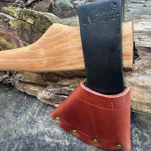 以下が含まれることがあります： 黒い頭と木製の柄が付いた斧で、「GENUINE Norlund」の文字が刻まれています。 斧の頭は、真鍮のリベットで固定された茶色の革製の鞘で覆われています。 斧は風化した木製の表面に置かれています。