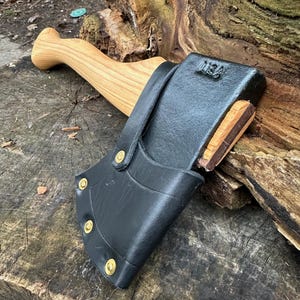 May include: A small axe with a wooden handle and a black leather sheath secured with brass rivets. The axe head is dark metal with a wooden wedge. The handle is light-colored wood. The axe is resting on a weathered wooden surface.