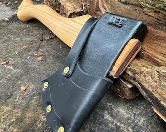 Puede incluir: Un hacha pequeña con mango de madera y funda de cuero negro asegurada con remaches de latón. La cabeza del hacha es de metal oscuro con una cuña de madera. El mango es de madera clara. El hacha descansa sobre una superficie de madera envejecida.
