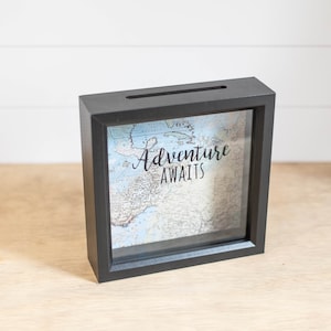 May include: Black wooden shadow box with a glass front. The box has a slot on the top for inserting money. Inside the box is a world map with the text "Adventure Awaits" printed on it.