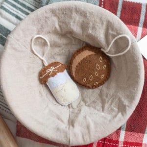May include: A beige linen bread proofing basket holds two felt ornaments: a cupcake and a cookie. The cupcake is brown, white, and blue, with a white bow. The cookie is brown with white seed details. The basket sits on a striped and plaid cloth.