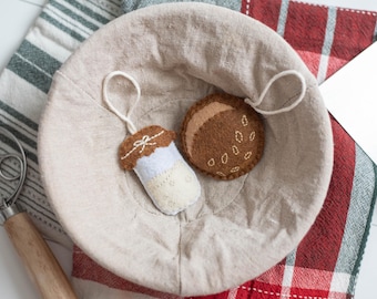 May include: A beige linen bread proofing basket holds two felt ornaments: a cupcake and a cookie. The cupcake is brown, white, and blue, with a white bow. The cookie is brown with white seed details. The basket sits on a striped and plaid cloth.