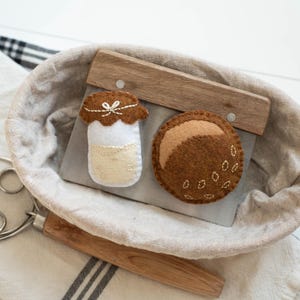 May include: A felt bread-making set includes a round loaf, a jar, and a bread scraper inside a linen-lined basket. The loaf is brown with seed details, and the jar has a brown lid. A dough whisk rests on a striped towel.