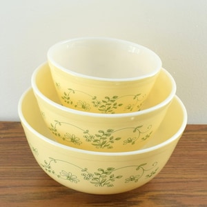 May include: A set of three nested mixing bowls in a pale yellow color, each featuring a delicate green floral vine pattern encircling the exterior. The bowls have a smooth, rounded shape with a wide rim and a clean, off-white interior finish.