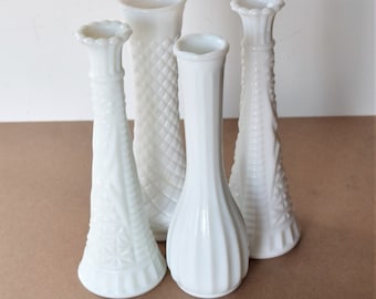 May include: Four white milk glass bud vases with different embossed patterns. The vases are arranged in a group on a brown surface.