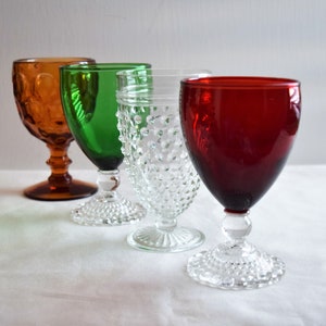 May include: Four vintage stemmed drinking glasses in different colors. The glasses are clear, green, amber, and red. The glasses have a textured, raised dot pattern.