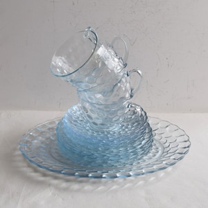 Puede incluir: Un conjunto de tazas y platillos de vidrio azul claro. Las tazas tienen asas y un patrón texturizado similar a burbujas. Los platillos están apilados debajo de las tazas, también con un patrón de burbujas. El conjunto está sobre una superficie blanca.