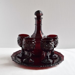 Puede incluir: Un juego de cuatro copas de vino de vidrio rojo rubí y una jarra a juego con un diseño decorativo. Las copas y la jarra están sobre una bandeja roja rubí a juego.