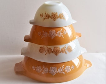Op de afbeelding: Een stapel van vier Pyrex mengkommen in wit en oranje. Elke kom heeft een bloemenpatroon in een contrasterende kleur. De kommen zijn genest, wat hun verschillende maten en klassieke ontwerp laat zien.