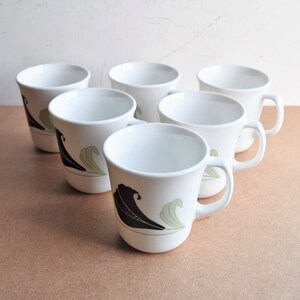 May include: Six white ceramic mugs with a black and green leaf design. Each mug has a handle and a curved shape. The mugs are arranged on a light brown surface.