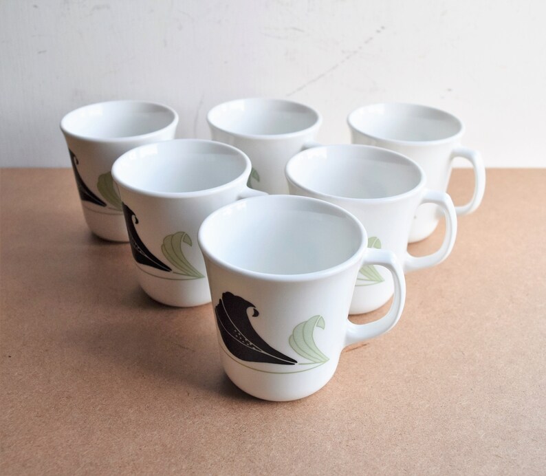 May include: Six white ceramic mugs with a black and green leaf design. Each mug has a handle and a curved shape. The mugs are arranged on a light brown surface.