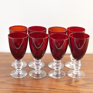 Puede incluir: Conjunto de ocho copas de vino vintage. Las copas presentan un cuenco rojo oscuro y un tallo transparente y decorativo. Los cuencos tienen forma redondeada y los tallos un diseño circular apilado. Las copas están sobre una superficie de madera.