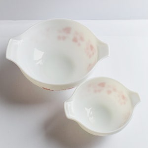 May include: Two white mixing bowls with pink floral designs. The bowls have handles on either side.