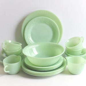 May include: A collection of jadeite green dishware, including plates, bowls, cups, and saucers. The plates and bowls have a ribbed design. The set is arranged on a white surface, showcasing the vintage style of the dinnerware.