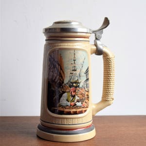 Op de afbeelding: Een keramische bierkan met een tinnen deksel en handvat. De kan heeft een kleurrijk beeld van een man die aan een schip werkt met een groot zeil op de achtergrond.