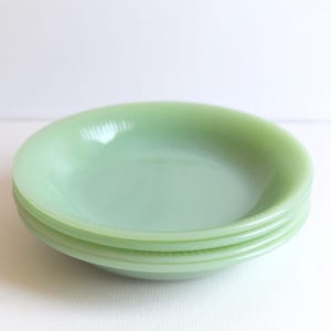 Puede incluir: Una pila de cuatro cuencos redondos poco profundos de color verde claro. Los cuencos tienen un acabado liso y brillante y un borde ligeramente texturizado. Los cuencos están apilados uno encima del otro, con el cuenco superior ligeramente inclinado.