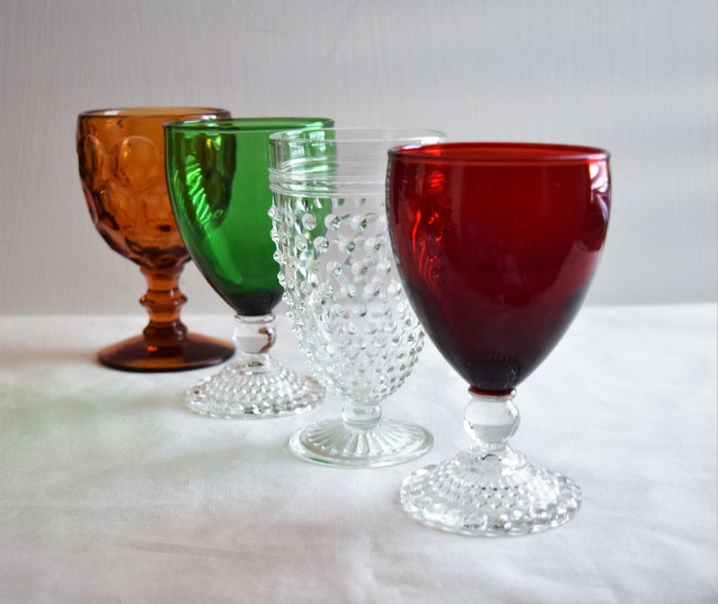 May include: Four vintage stemmed drinking glasses in different colors. The glasses are clear, green, amber, and red. The glasses have a textured, raised dot pattern.