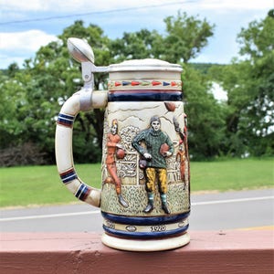 May include: A ceramic beer stein featuring a relief design of football players in vintage uniforms. The handle is topped with a metal football-shaped lid finial. The base includes the years 1900 and 1920 printed in white on a dark blue band.