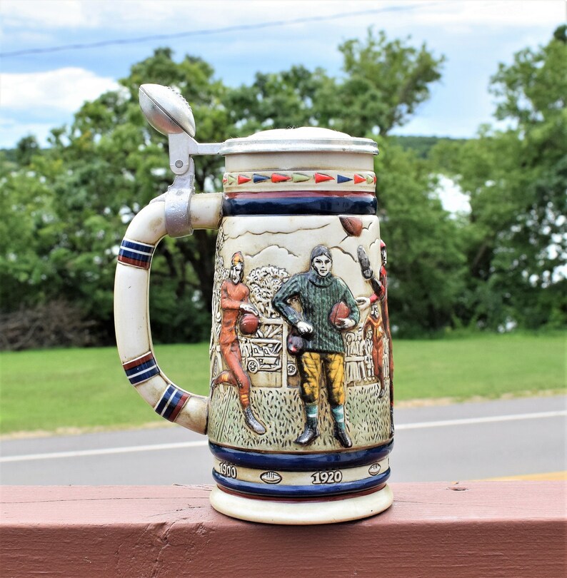 May include: A ceramic beer stein featuring a relief design of football players in vintage uniforms. The handle is topped with a metal football-shaped lid finial. The base includes the years 1900 and 1920 printed in white on a dark blue band.