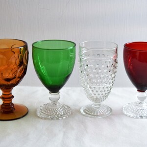 Op de afbeelding: Vier wijnglazen met steel in verschillende kleuren en patronen. De glazen zijn van links naar rechts gerangschikt: amberkleurig met een bubbelpatroon, groen, helder met een bubbelpatroon en rood.