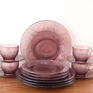 Pode incluir: Um conjunto de jantar de vidro lavanda vintage. O conjunto inclui pratos, pires e ch&aacute;venas de ch&aacute;. Os pratos e ch&aacute;venas apresentam um design floral em relevo. O conjunto est&aacute; disposto numa superf&iacute;cie de madeira sobre um fundo branco.