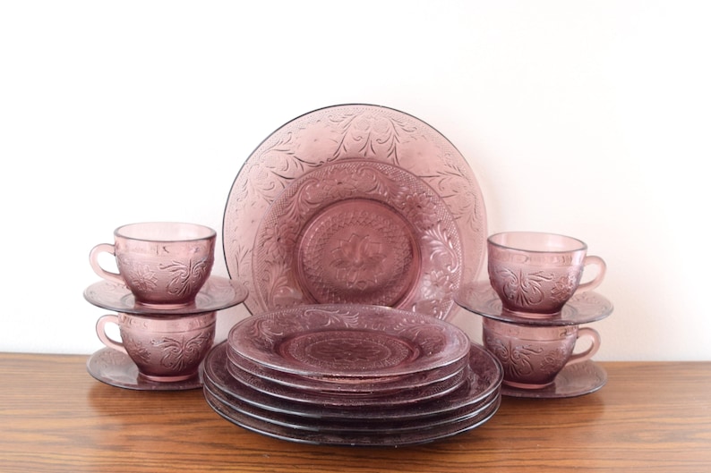 Pode incluir: Um conjunto de jantar de vidro lavanda vintage. O conjunto inclui pratos, pires e ch&aacute;venas de ch&aacute;. Os pratos e ch&aacute;venas apresentam um design floral em relevo. O conjunto est&aacute; disposto numa superf&iacute;cie de madeira sobre um fundo branco.