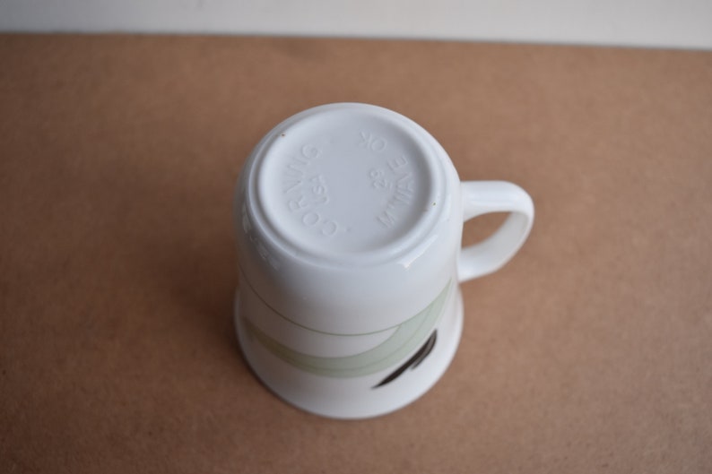 May include: White ceramic mug with a curved handle, featuring a design of green and black curved lines. The bottom of the mug has the text "CO. RIWING USA 29 WAVE OK".