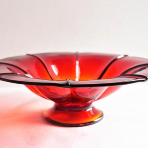 May include: A red glass bowl with a fluted rim and a pedestal base. The bowl is made of clear glass with a red tint.