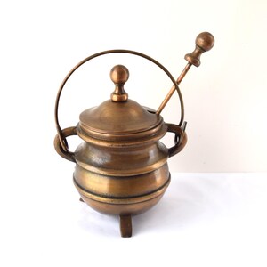 May include: A round, metallic bronze-toned pot with three short legs, two side handles, and a matching lid with a central knob. A thin, arched metal handle extends over the top, and a long, slender spoon with a spherical finial rests inside.