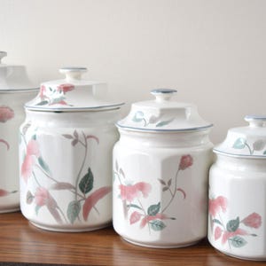 Puede incluir: Juego de cuatro botes de cerámica blanca con tapas. Cada bote presenta un diseño floral con detalles en rosa y verde. Los botes están dispuestos en fila sobre una superficie de madera, de diferentes tamaños.