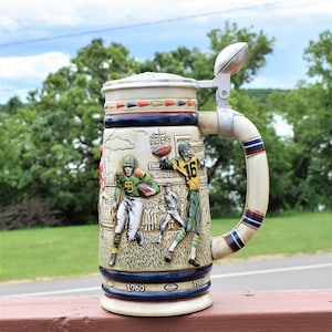 May include: A ceramic beer stein featuring a relief design of football players in action on a field. The stein has a metal lid with a football-shaped finial, a curved handle with blue and red stripes, and the years 1960 and 1980 printed at the base.