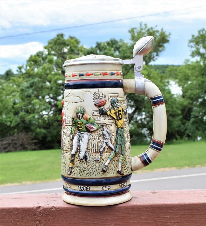May include: A ceramic beer stein featuring a relief design of football players in action on a field. The stein has a metal lid with a football-shaped finial, a curved handle with blue and red stripes, and the years 1960 and 1980 printed at the base.
