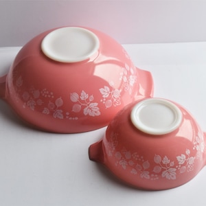 May include: Two pink Pyrex mixing bowls with a white gooseberry pattern. The bowls are in different sizes and have a white interior.