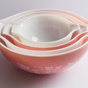 May include: Set of four pink nesting mixing bowls with white rims and a white floral pattern. The bowls are made of glass and have a vintage look.