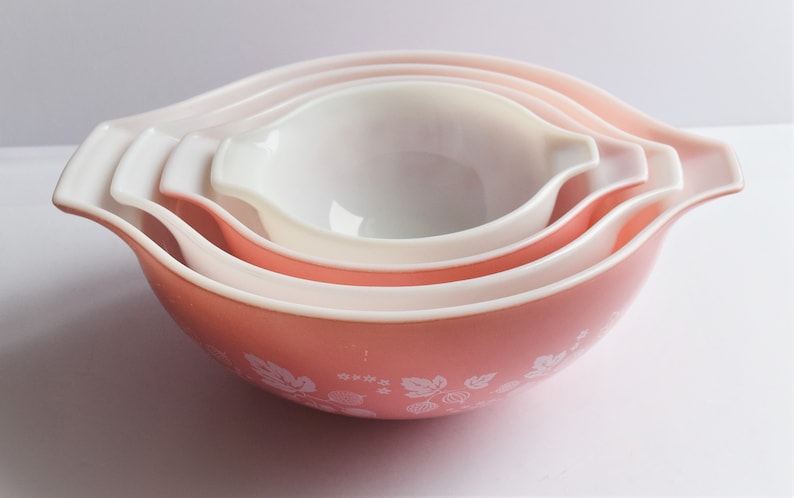 May include: Set of four pink nesting mixing bowls with white rims and a white floral pattern. The bowls are made of glass and have a vintage look.