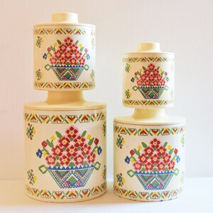 May include: Set of three white ceramic canisters with floral designs. The canisters have a cream base with a colorful floral pattern and a geometric border.