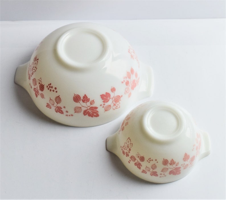 May include: Two white ceramic bowls with pink gooseberry designs. The bowls have handles on either side.
