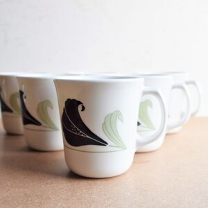 May include: Set of six white ceramic mugs. Each mug features a black and green floral design. The mugs have a curved shape with a handle on the side. The background is a light tan color.