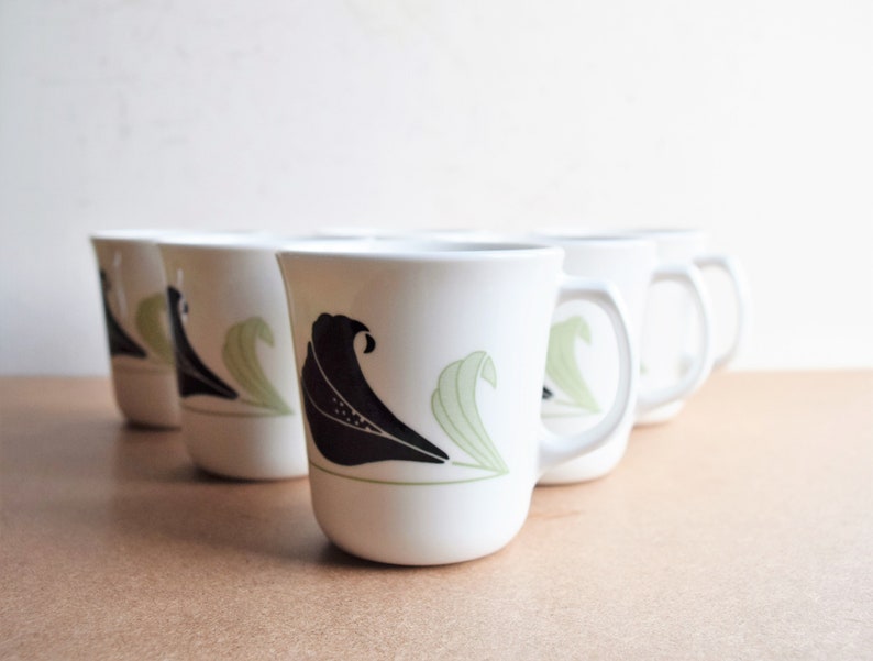 May include: Set of six white ceramic mugs. Each mug features a black and green floral design. The mugs have a curved shape with a handle on the side. The background is a light tan color.