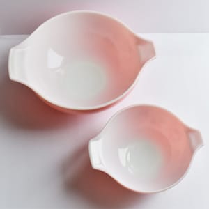 May include: Two pink glass mixing bowls with handles. The larger bowl is a lighter shade of pink than the smaller bowl.